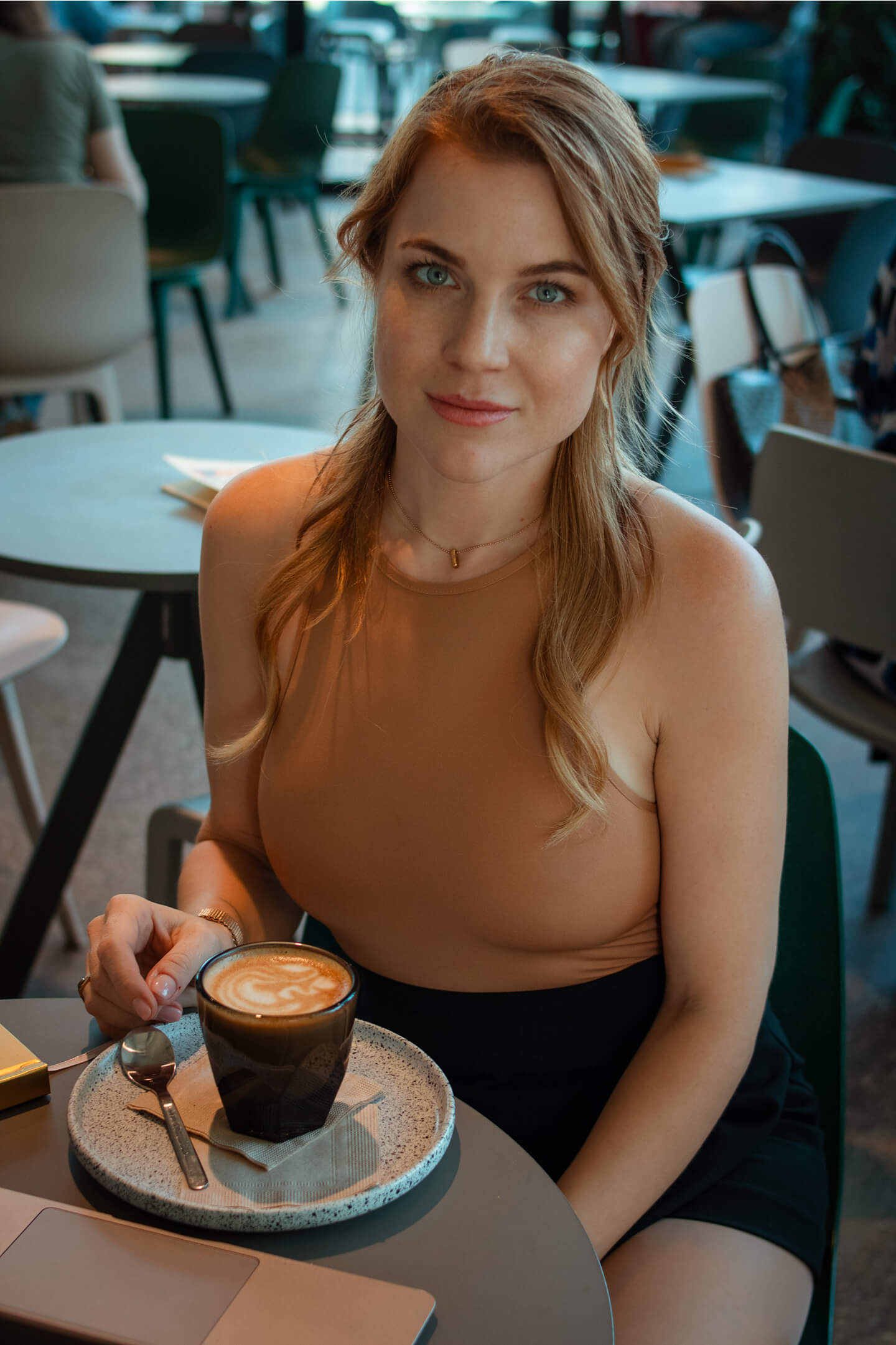 Nő barna felsőben ül cappuccinóval a kávézói asztalnál