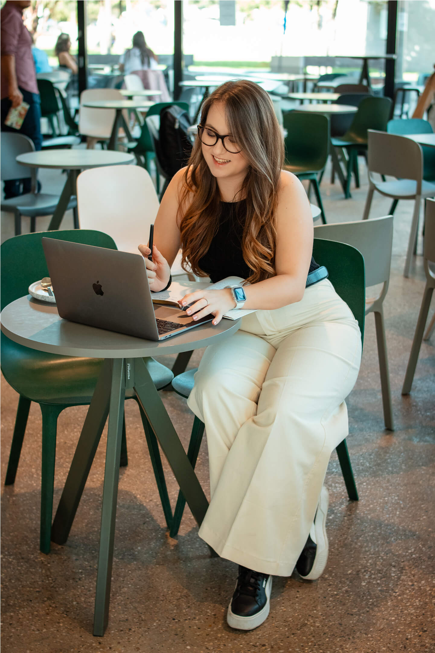 Fiatal nő ül mosolyogva a laptopja mögött, kávézóban dolgozik