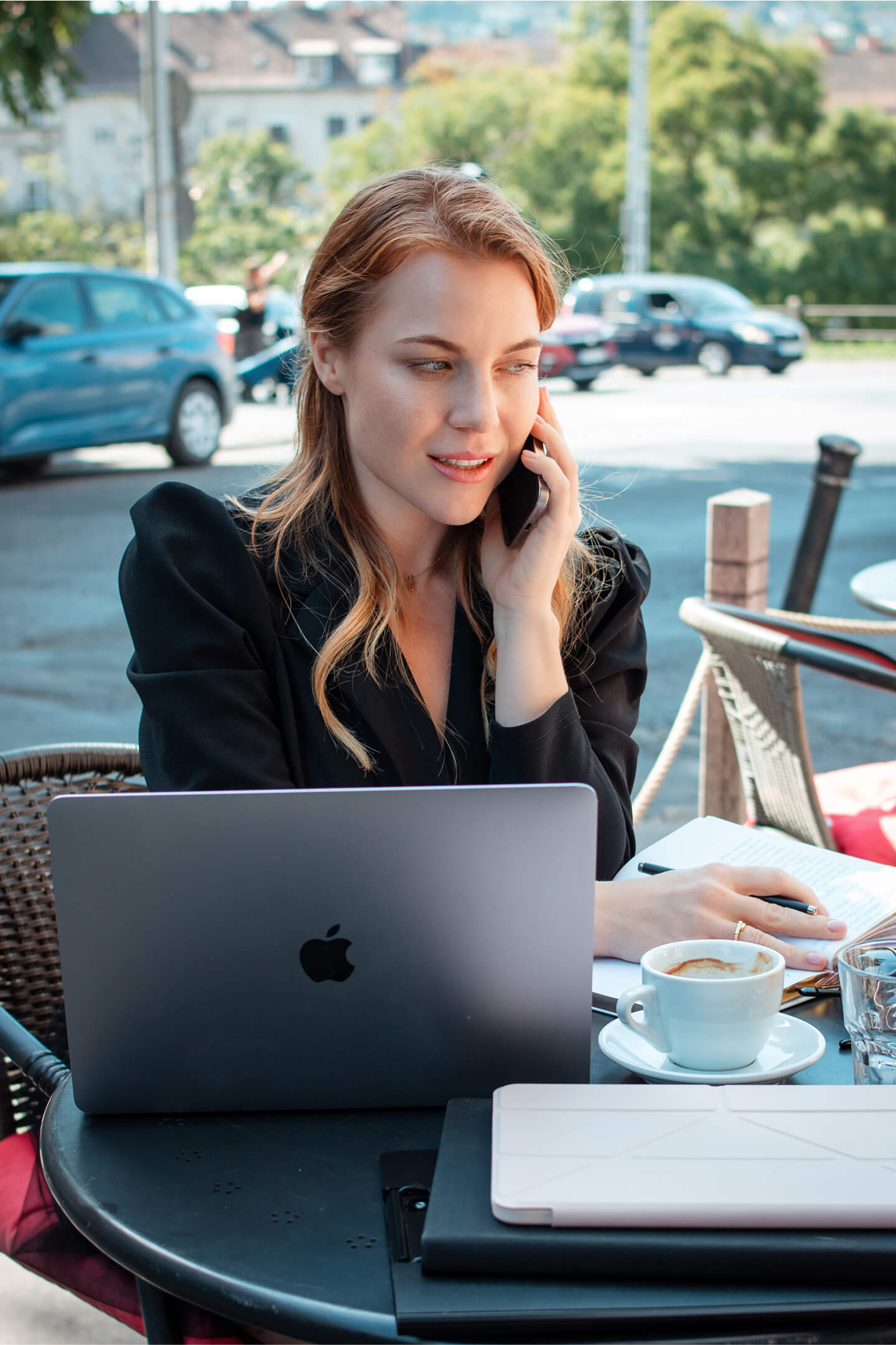 Nő fekete zakóban ül a laptopja előtt, telefonál, cappuccinóval