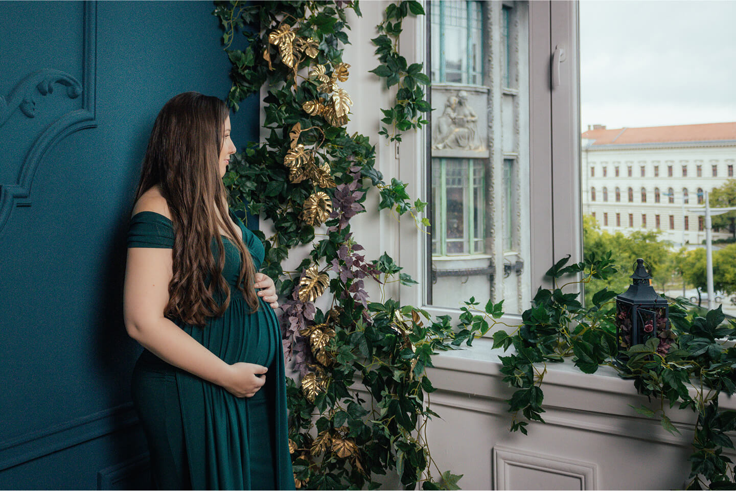 Kismama zöld ruhában az ablaknál állva, növényekkel körülvéve