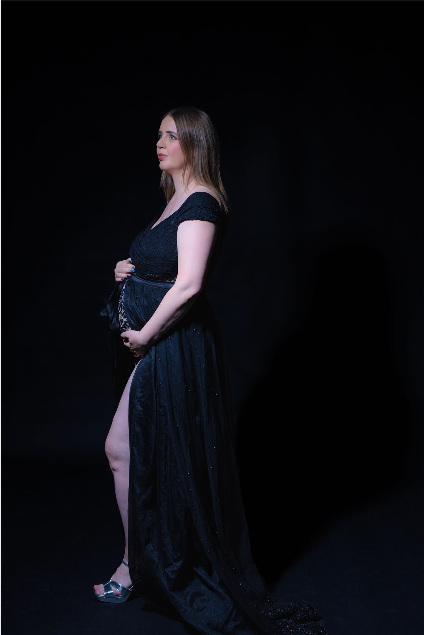 Kismama oldalt állva, csipkeruhában, stúdiófényekkel megvilágítva.