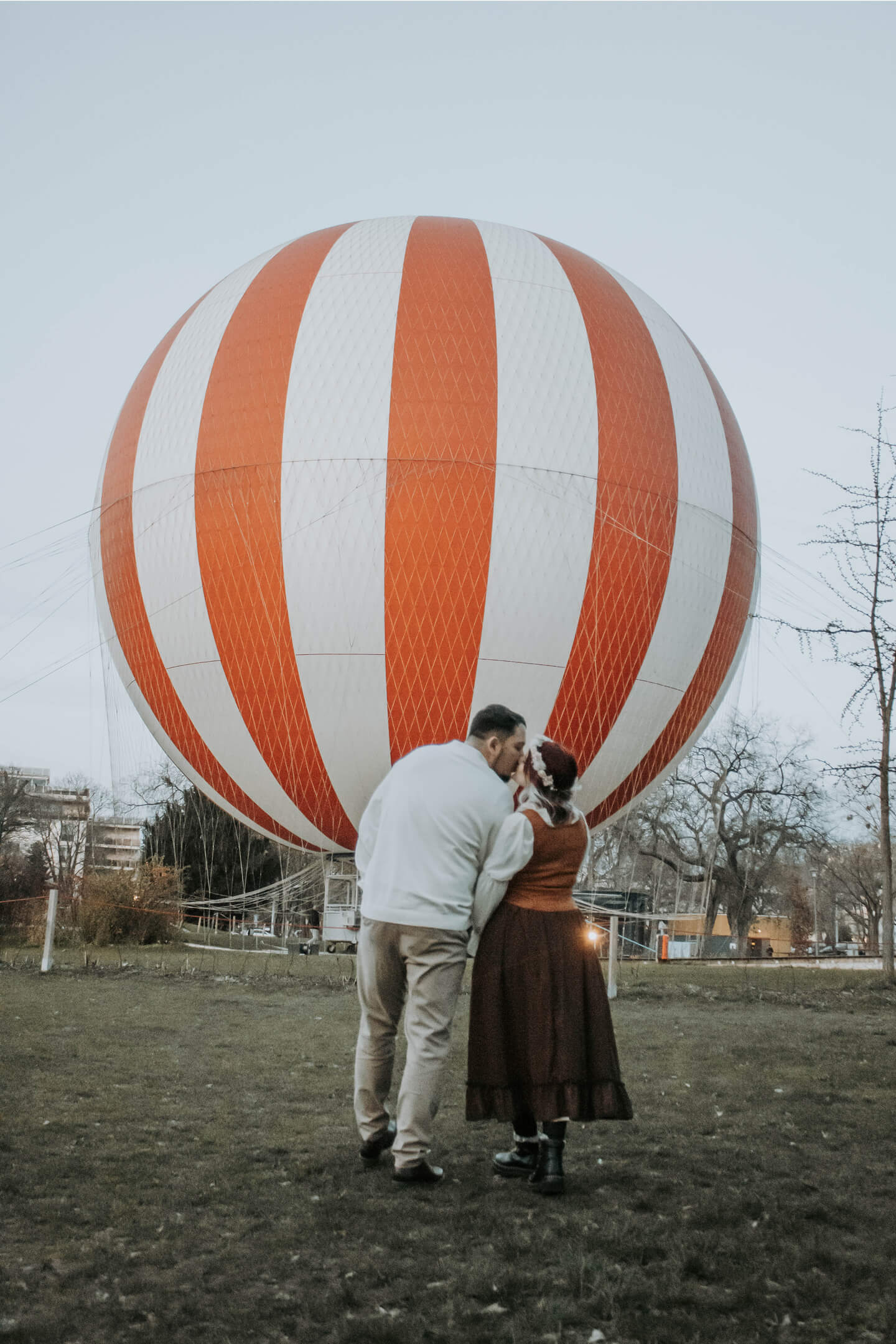Jegyespár csókja egy hatalmas narancs-fehér hőlégballon előtt.