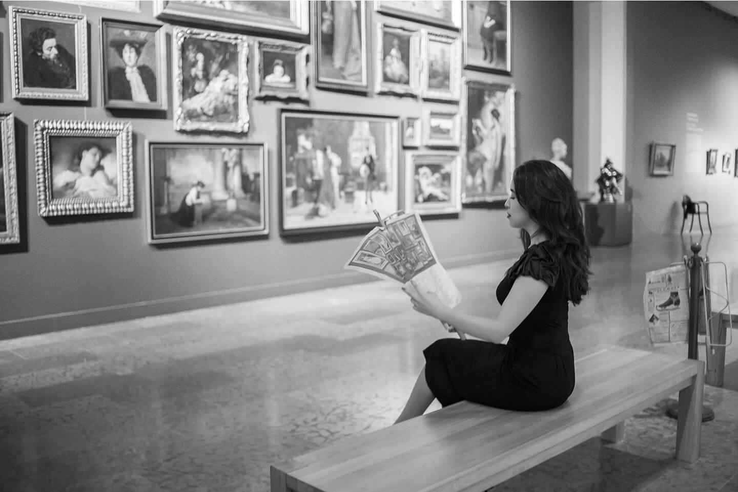 Woman sitting on a bench reading a vintage newspaper in an art gallery.